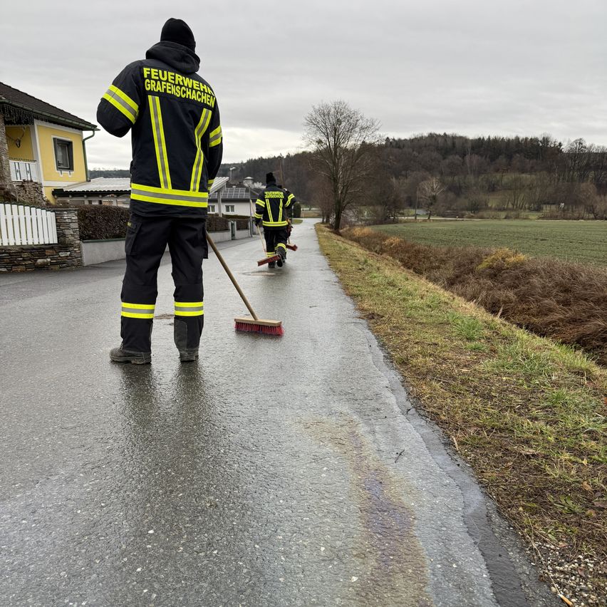 Zwei Feuerwehrleute reinigen einen Pfad mit Besen. Einer steht, während der andere geht. Sie tragen reflektierende Jacken mit 'Feuerwehr' und 'Grafenschachen' darauf. Hinter ihnen steht ein gelbes Haus mit einem weißen Zaun.