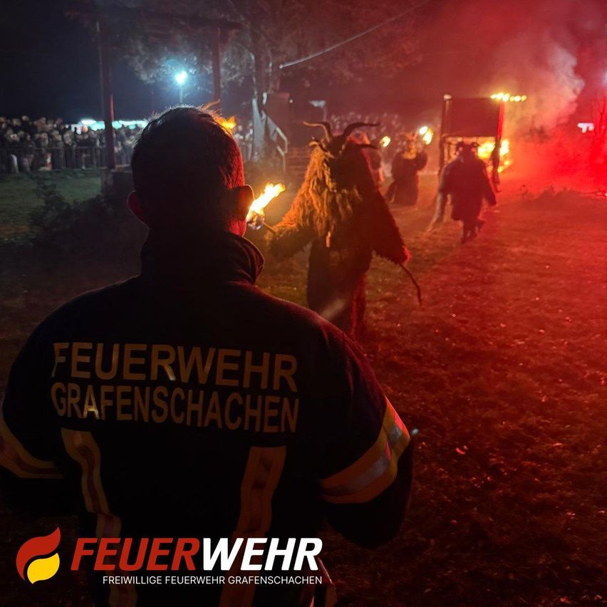 Ein Feuerwehrmann beobachtet von hinten, wie Menschen in Kostümen mit Hörnern und Fackeln vorbeigehen. Der Boden ist rot beleuchtet.