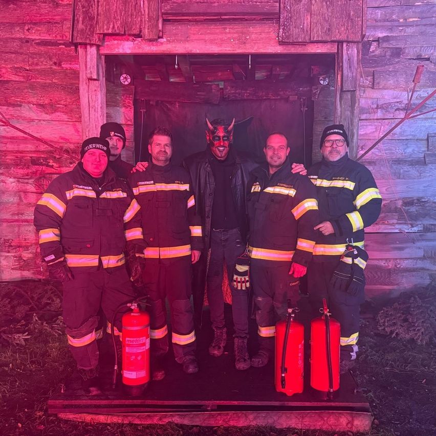 Fünf Feuerwehrleute stehen vor einer Holzhütte, einer trägt eine rote Maske und einer hält einen Feuerlöscher. Sie alle haben feuerfeste Uniformen an.