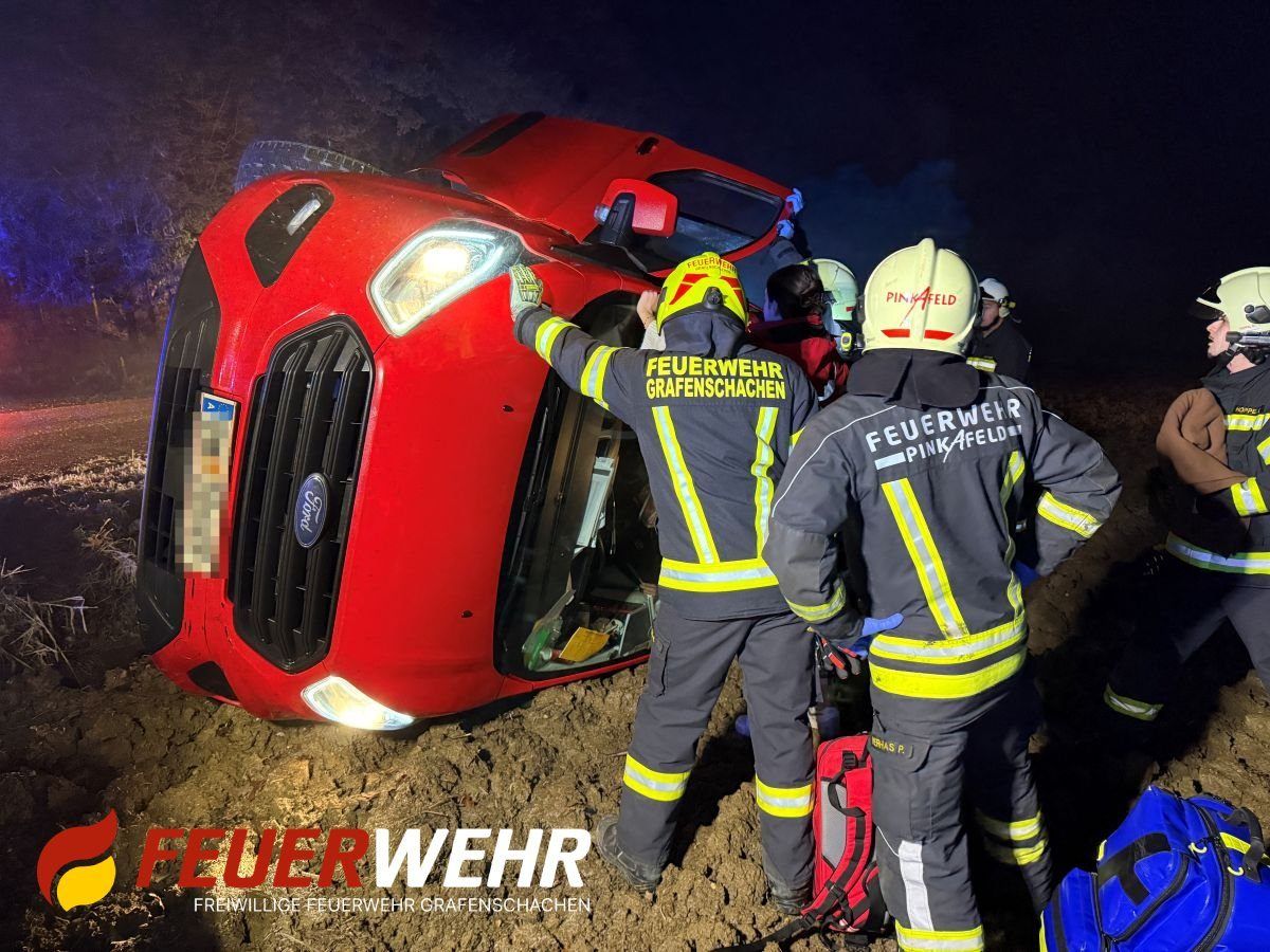 Feuerwehrleute kümmern sich um ein rotes Auto, das auf einer schlammigen Straße nachts auf der Seite liegt. Zwei Feuerwehrleute unterstützen eine Person in der Nähe des Fahrzeugs, während andere in der Nähe stehen.