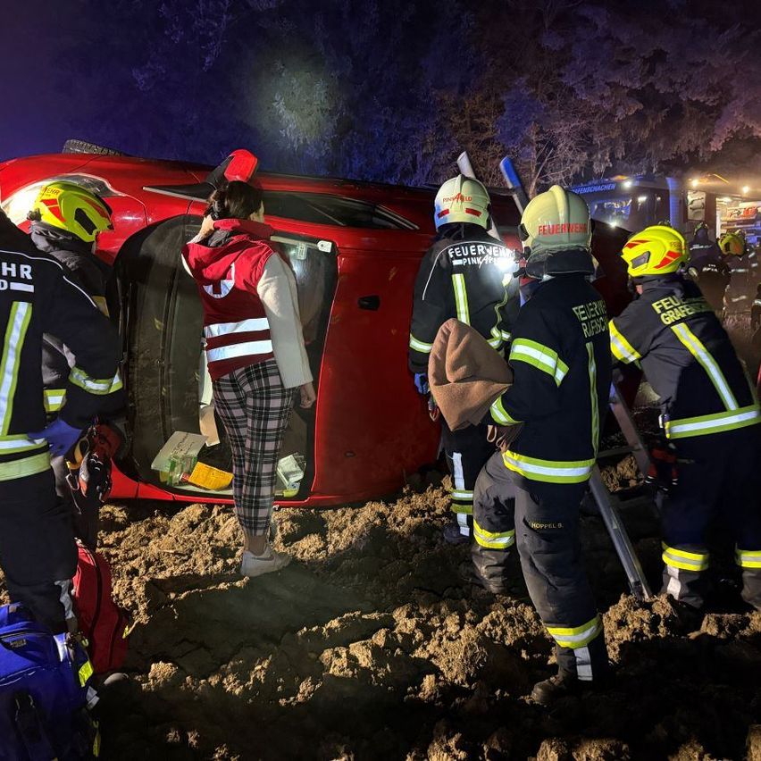 Feuerwehrleute helfen einer Frau aus einem roten Auto auf einem schlammigen Weg bei Nacht. Das Auto ist beschädigt.