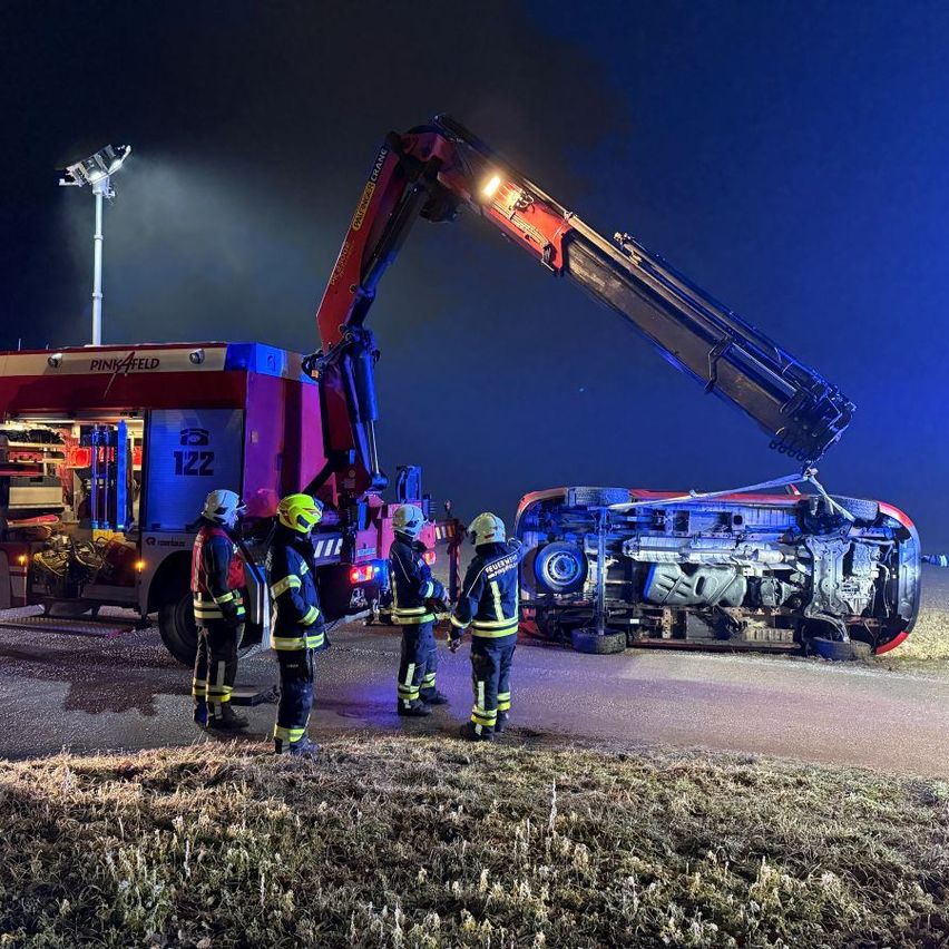 Ein Feuerwehrauto steht mit einem Kran, der ein beschädigtes Auto hebt, auf der Straße. Fünf Feuerwehrleute stehen in der Nähe, und eine Straßenlaterne steht hinter dem Feuerwehrauto.