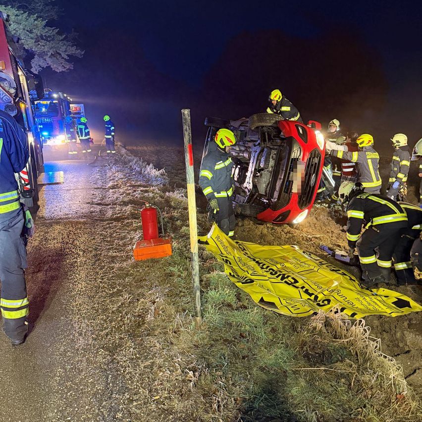 Feuerwehrleute in gelben Helmen arbeiten an einem umgekippten roten Auto. Ein gelber Teppich ist auf dem Boden ausgebreitet.