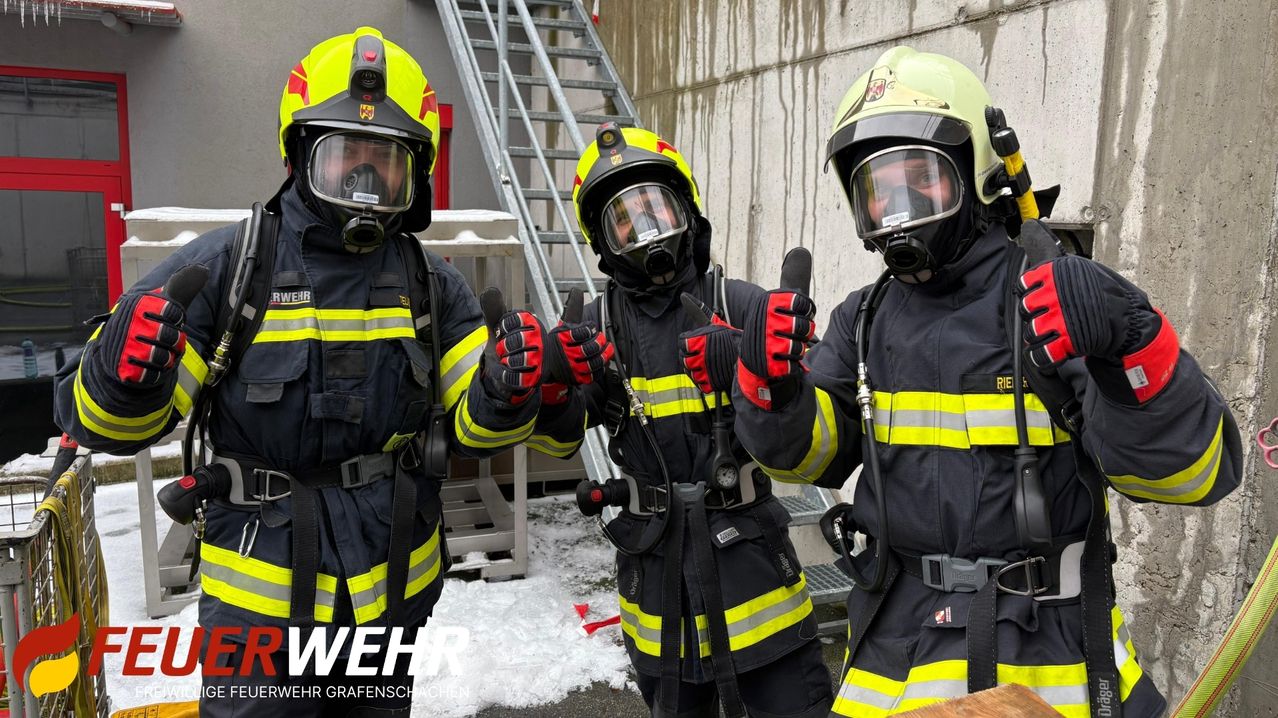 Three firefighters in full gear, including helmets and gas masks, are giving thumbs up in front of a concrete wall with a staircase and snow on the ground.