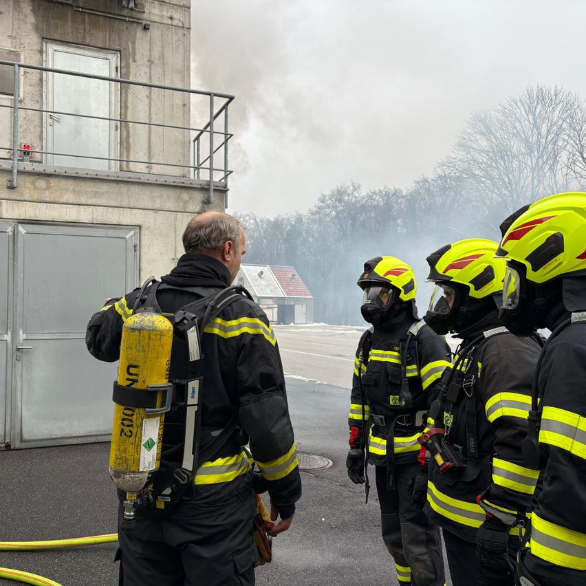 Firefighters in yellow and black uniforms with helmets are gathered, possibly preparing for a fire. A firefighter with a yellow oxygen tank is looking to the side. Smoke rises from the area behind them.
