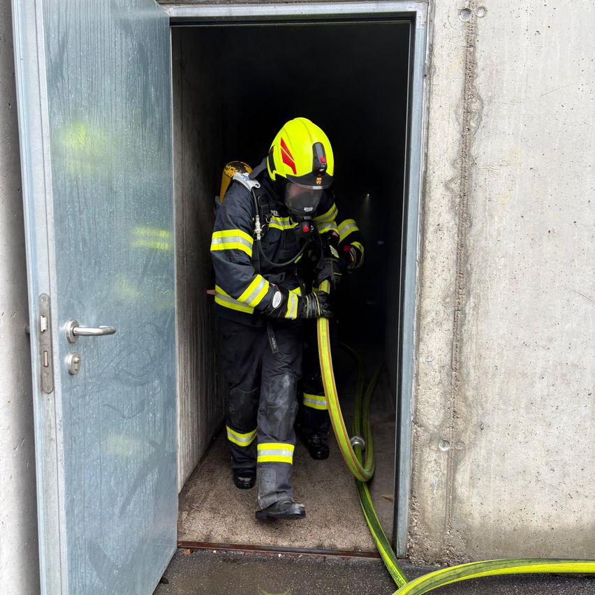 A firefighter wearing a yellow helmet and full protective gear stands inside a building, holding a hose.