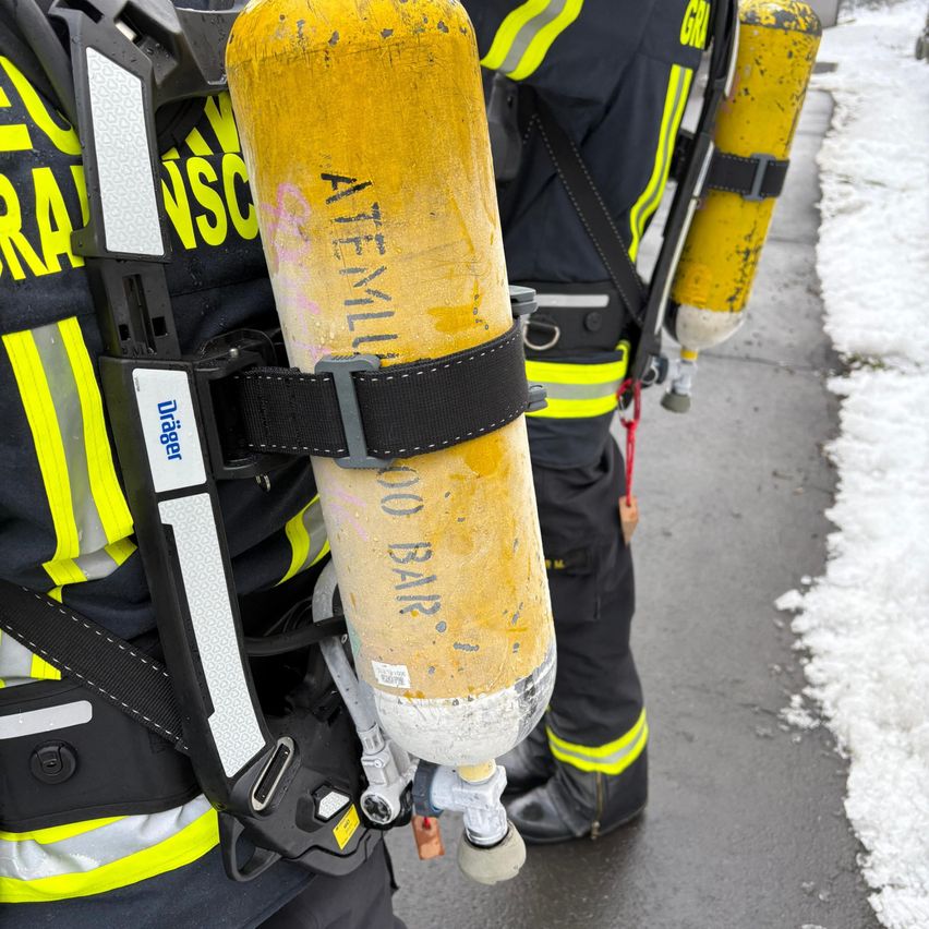 Two firefighters in protective gear are walking on a snowy street, one carrying a yellow oxygen tank labeled Atemiulu.