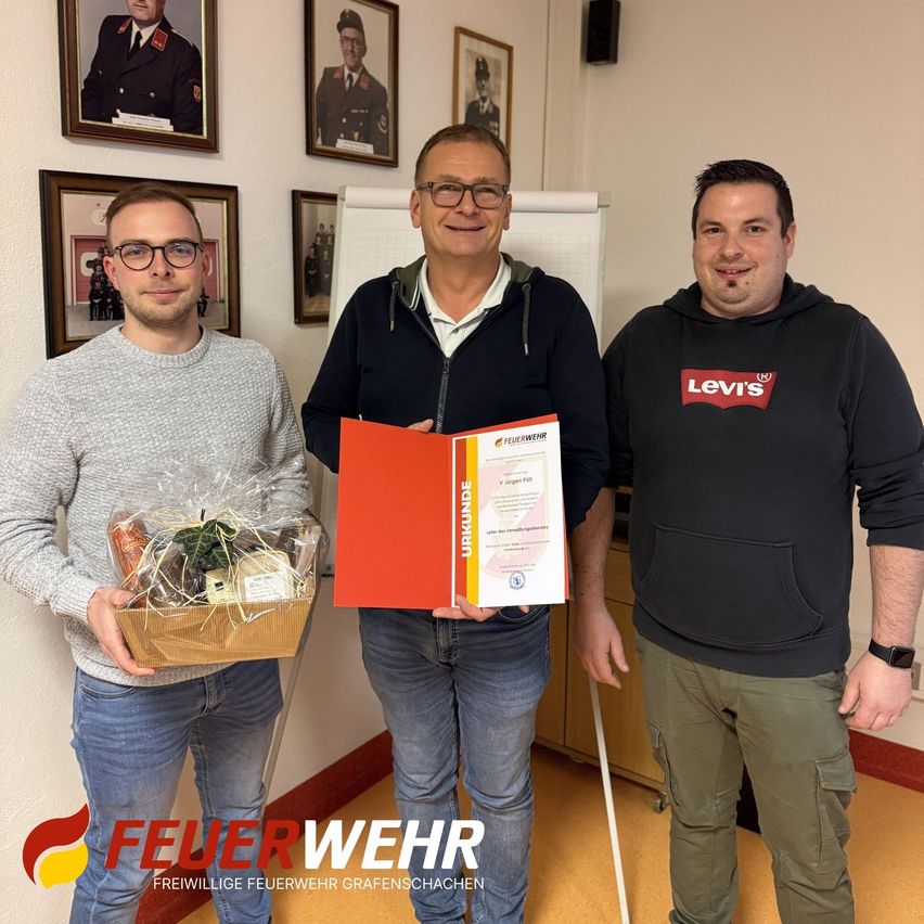 Drei Männer stehen in einem Raum mit gerahmten Bildern an der Wand. Der Mann auf der linken Seite hält einen Geschenkkorb. Der Mann in der Mitte hält ein Zertifikat und lächelt. Der Mann auf der rechten Seite trägt eine Levi's Kapuzenjacke.