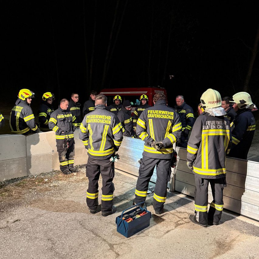 A group of firefighters in uniform are standing in front of a concrete wall at night. They are probably preparing for an emergency.