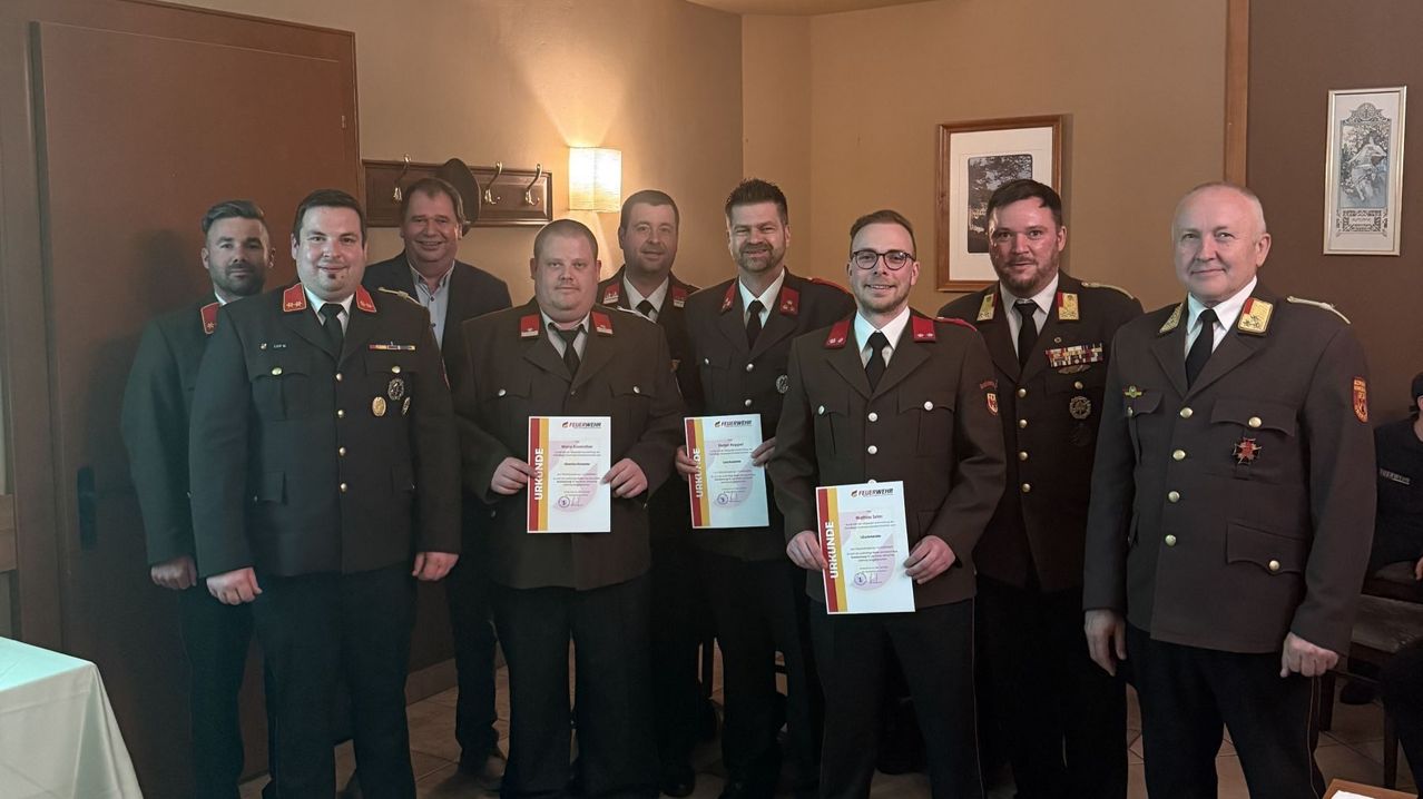 Eine Gruppe uniformierter Männer posiert mit Zertifikaten für ein Foto. Sie stehen in einem Raum mit warmer Beleuchtung.