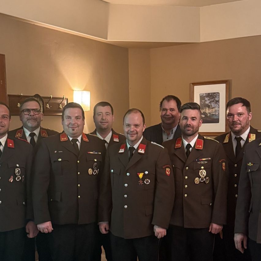 Eine Gruppe von Männern in Militäruniformen steht in einem Raum und lächelt für ein Foto. An der Wand hinter ihnen befindet sich ein Bilderrahmen.