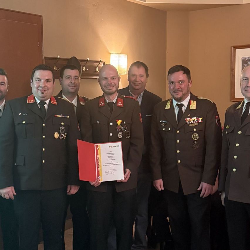 Mehrere Militäroffiziere stehen in einem Raum zusammen. Die zentrale Figur hält ein Zertifikat in der Hand und trägt eine Medaille.