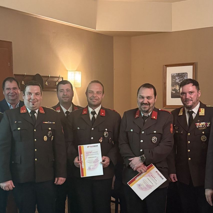 Eine Gruppe von Männern in Militäruniformen posiert für ein Foto, einer hält ein Zertifikat. Sie stehen in einem Raum mit einem gerahmten Bild an der Wand.