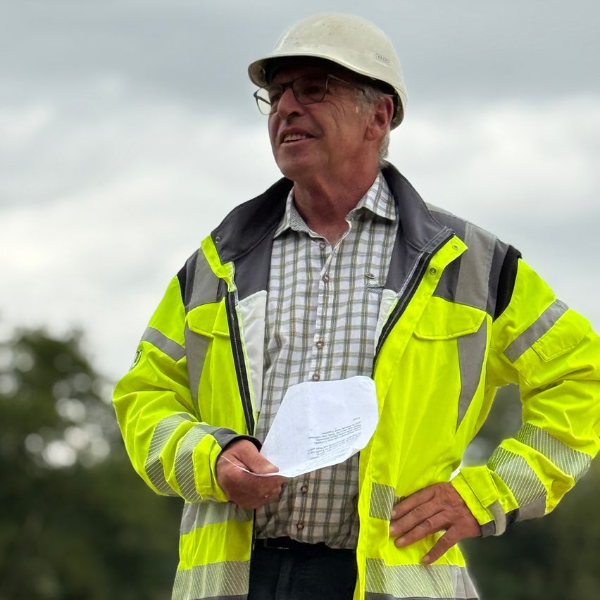 An older man wearing a reflective safety jacket and a white hard hat is standing outdoors, holding a piece of paper and smiling.