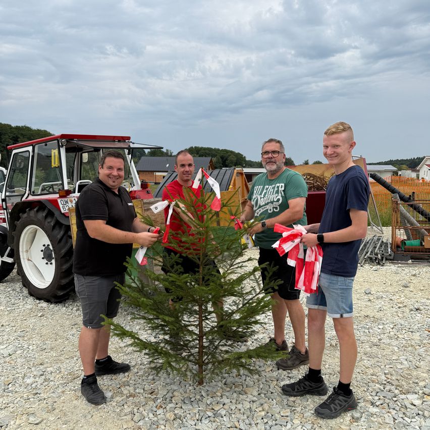 Four men stand around a small tree, holding red and white ribbons, possibly celebrating a planting event. Behind them is a tractor and a cloudy sky.
