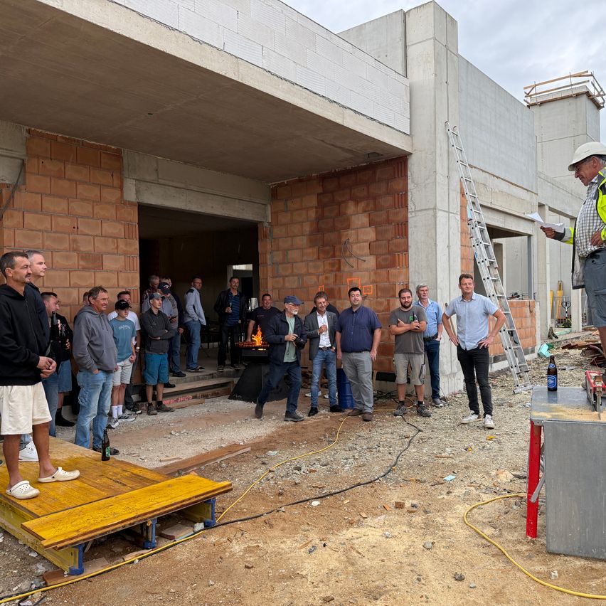 A group of people stand in front of a building under construction, with a man on a platform holding a piece of paper. A fire pit is visible inside the building.