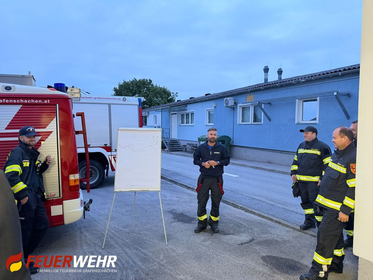 A firefighter stands in front of a whiteboard, with another firefighter standing behind him. They are outside near a fire truck and a blue building.