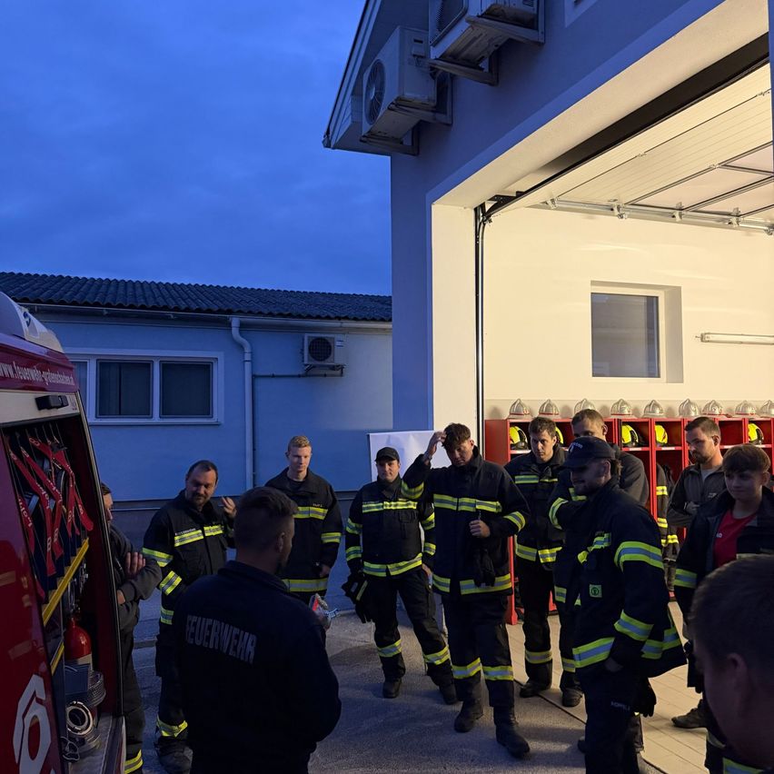 A group of firefighters in uniform are gathered outside a fire station at night, with some standing by a fire truck.