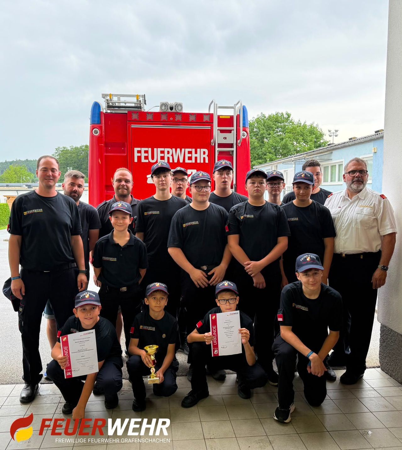 Eine Gruppe von Feuerwehrleuten posiert für ein Foto mit einem roten Feuerwehrauto im Hintergrund. Sie halten Urkunden und einen Pokal in der Hand, gekleidet in Uniformen und Mützen.
