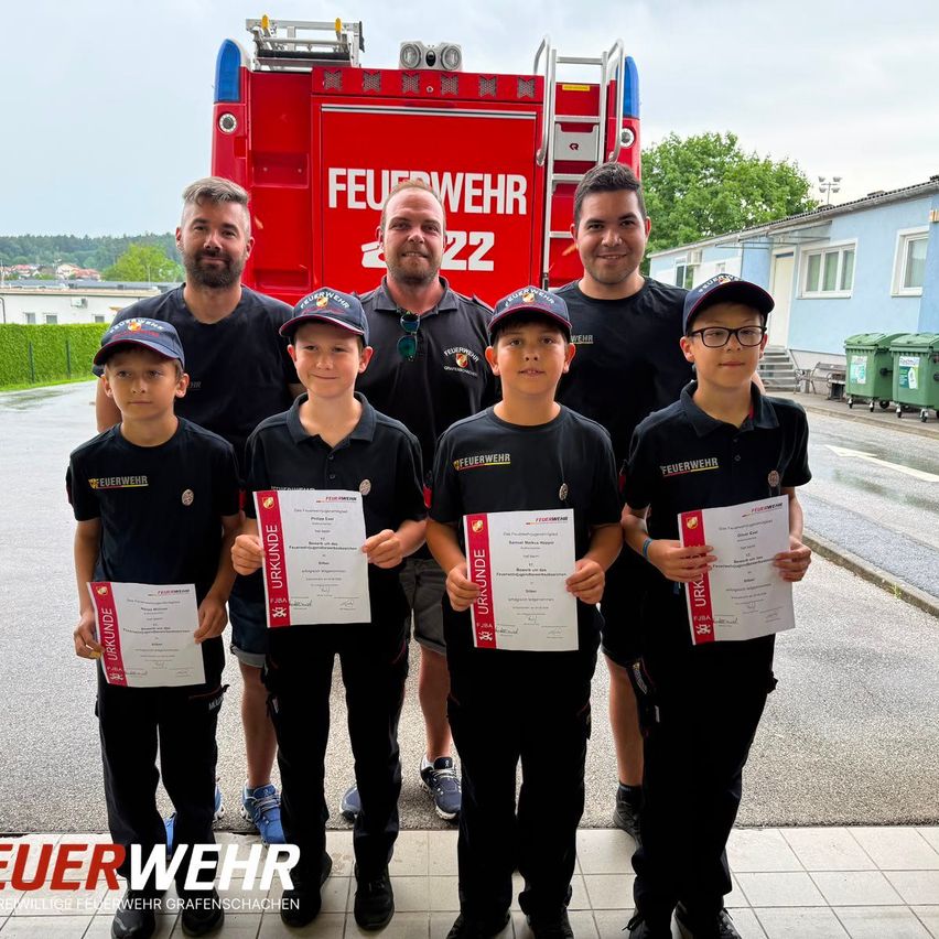Fünf Jungen in Uniformen halten Urkunden vor einem Feuerwehrauto mit zwei erwachsenen Männern. Sie stehen draußen mit einem blauen Gebäude und grünen Mülleimern im Hintergrund.
