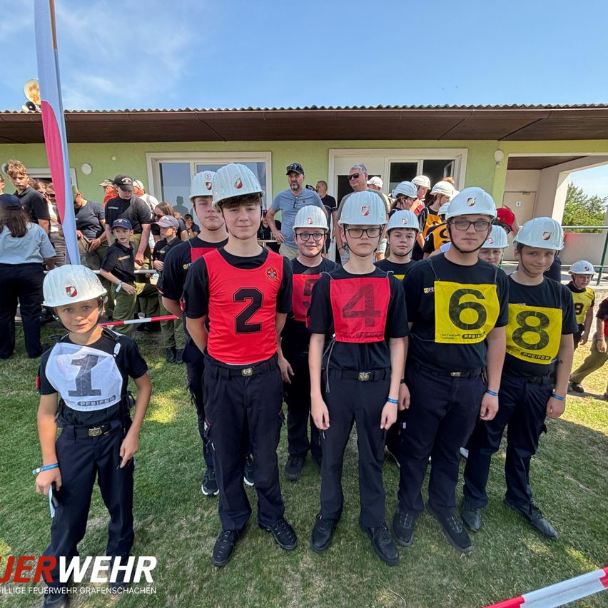 Eine Gruppe von Kindern und Erwachsenen in Helmen und nummerierten Uniformen steht auf einem Rasenfeld vor einem Gebäude.
