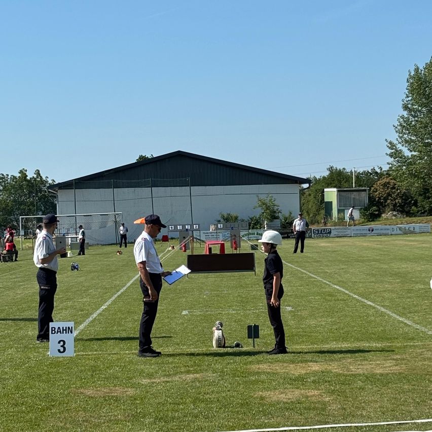 Drei Beamte stehen auf einem Feld mit einem Schild, das Bahn 3 beschriftet ist. Ein Junge mit Helm beobachtet sie, und ein Hund ruht in der Nähe. Ein Gebäude mit Zaun und mehrere Personen im Hintergrund.