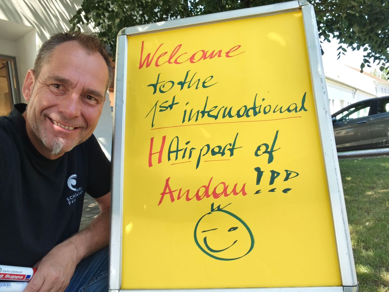 Ein Mann lächelt neben einem gelben Schild, auf dem steht Willkommen am 1. internationalen HAirport von Andou!!