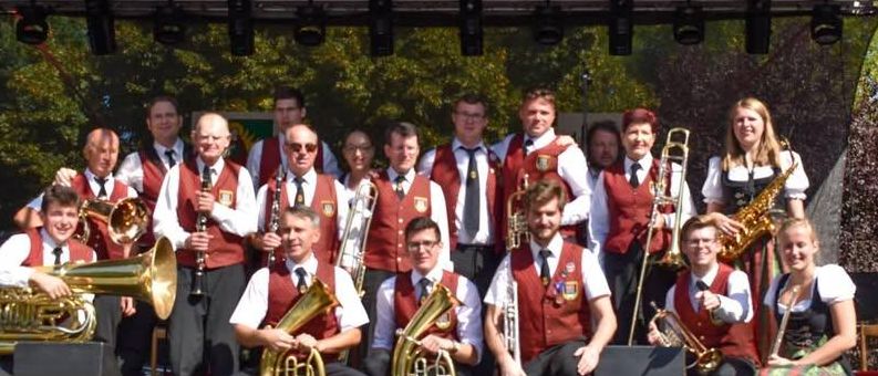 Ein Blasorchester posiert für ein Gruppenfoto auf einer Bühne mit Vordach, hält verschiedene Musikinstrumente und trägt passende rote Westen.