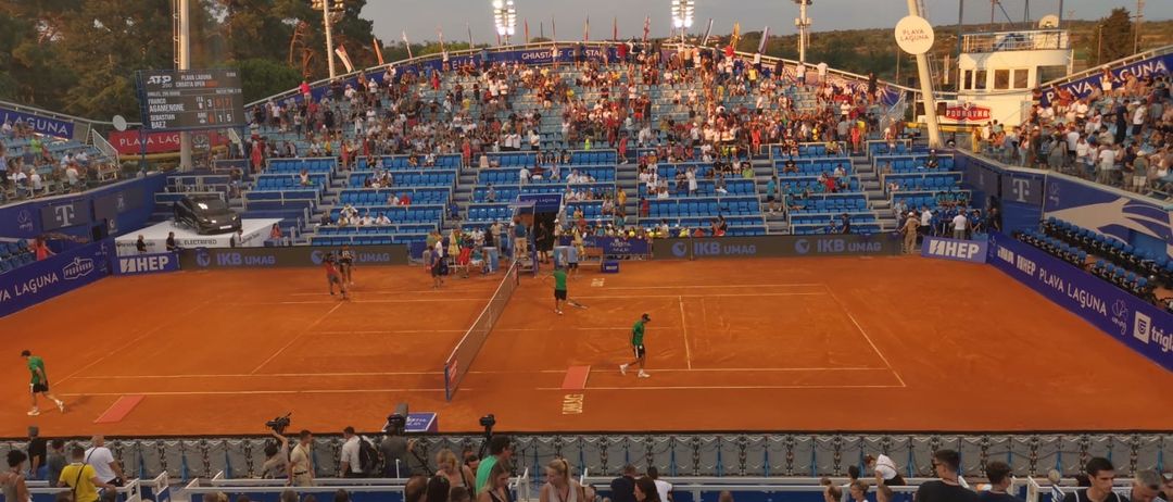 Ein Tennismatch findet in einem Stadion statt, mit Spielern in Grün und Weiß. Die Zuschauer füllen die blauen Tribünen und schauen aufmerksam zu. Helle Lichter beleuchten den Platz, mit Werbetafeln und Flaggen sichtbar.