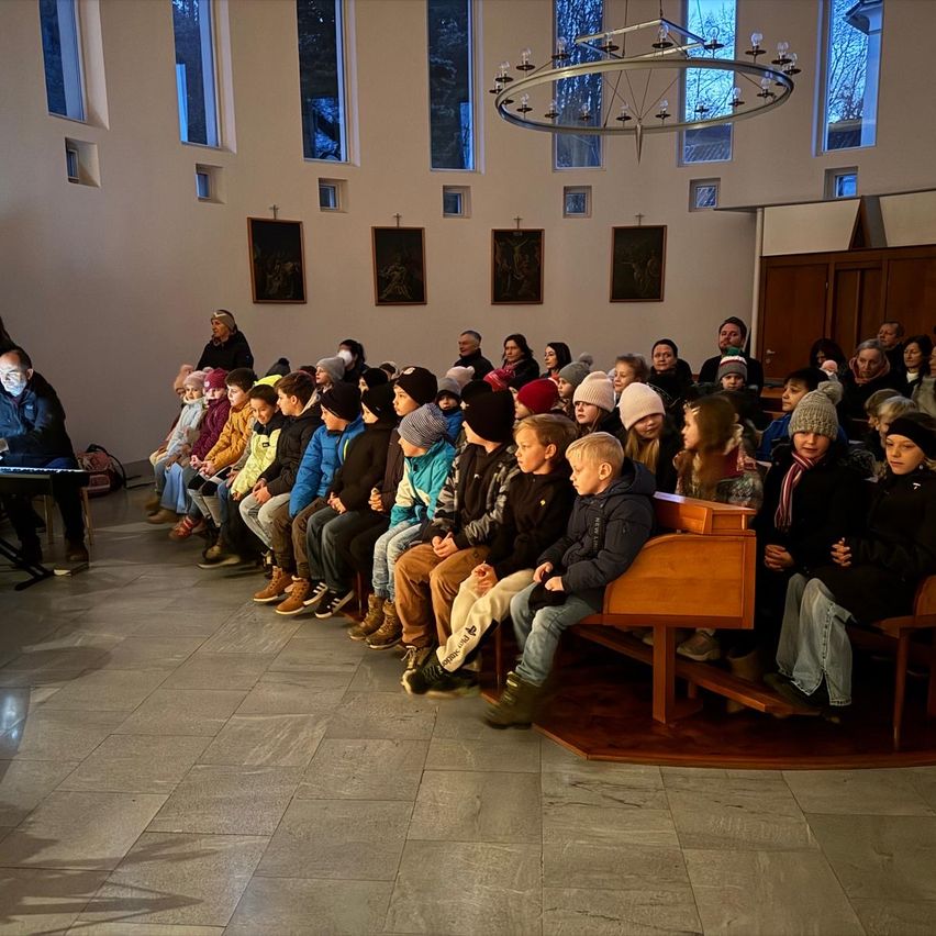 Eine Gruppe von Kindern und Erwachsenen sitzt in einer Kirche, vor einem Pianisten. Die Kirche hat gewölbte Fenster und einen Kronleuchter. Alle tragen Winterkleidung.