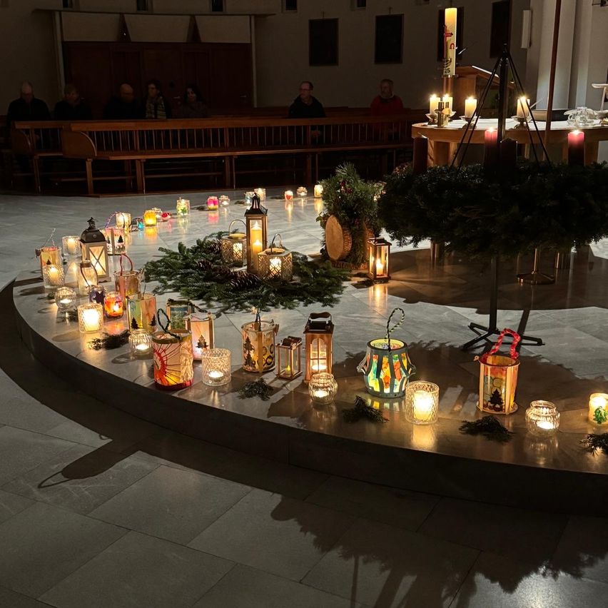 Eine kreisförmige Anordnung von brennenden Laternen und Kerzen auf einem gefliesten Boden in einer Kirche, mit Menschen im Hintergrund, die sitzen.