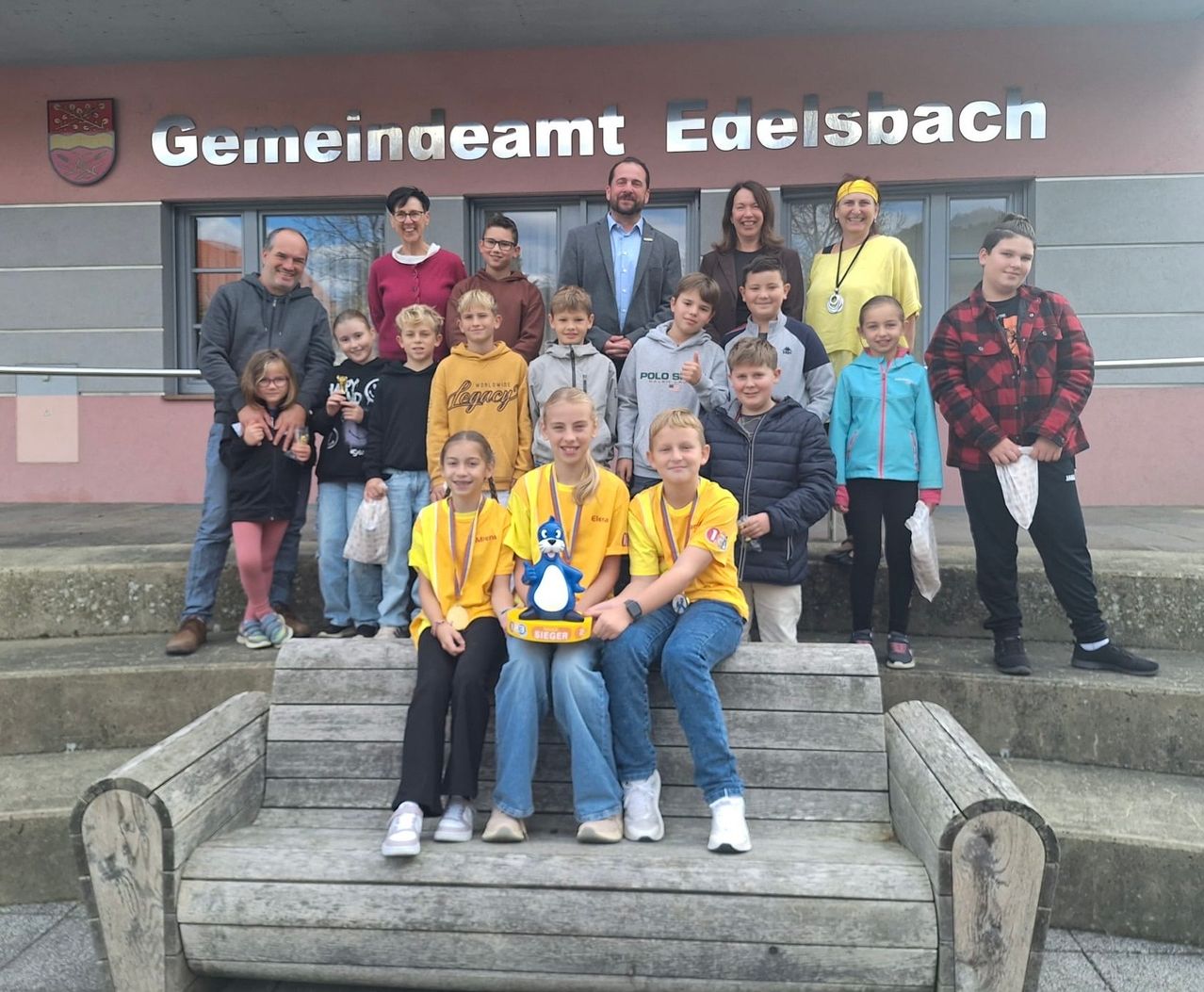 Eine Gruppe von Kindern und Erwachsenen posiert für ein Foto vor dem Gemeindeamt Eeielspach. Die Kinder tragen gelbe T-Shirts mit Medaillen, und Erwachsene stehen hinter ihnen.