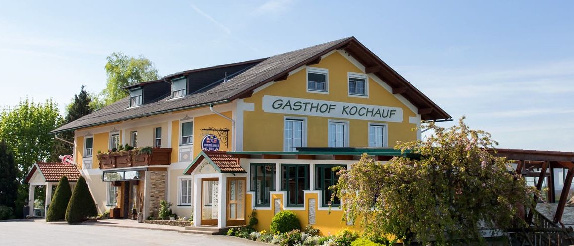 Der Gasthof Kochauf ist ein gelbes Gebäude mit mehreren Fenstern, einem roten Dach und Pflanzen draußen. Das Schild sagt Gasthof Kochauf.