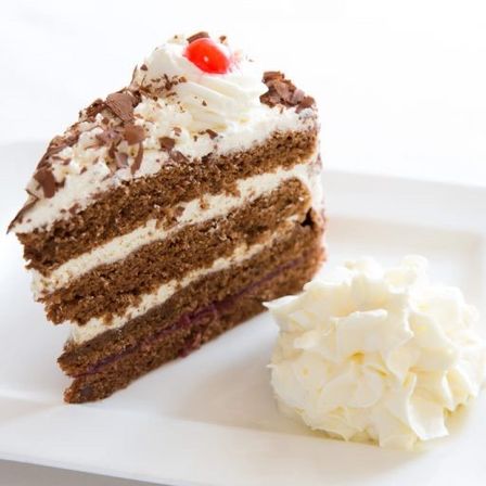 Ein Stück Kuchen mit weißer und Schokoladenglasur, gekrönt mit Schlagsahne und einer Kirsche, auf einem weißen Teller.