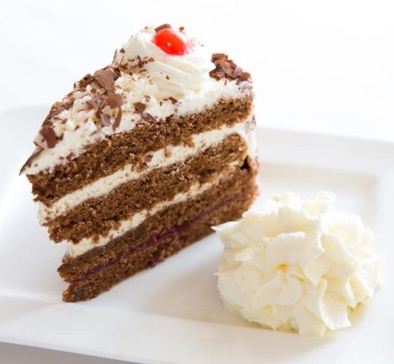 Ein Stück Kuchen mit weißer und Schokoladenglasur, gekrönt mit Schlagsahne und einer Kirsche, auf einem weißen Teller.