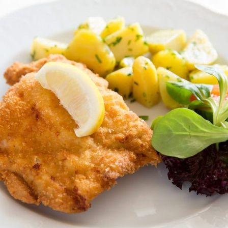 Auf einem weißen Teller befindet sich ein Stück gebratenes Fleisch mit einer Zitronenscheibe, neben geschnittenen Kartoffeln und einem grünen Salat.