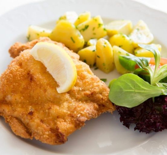 Auf einem weißen Teller befindet sich ein Stück gebratenes Fleisch mit einer Zitronenscheibe, neben geschnittenen Kartoffeln und einem grünen Salat.