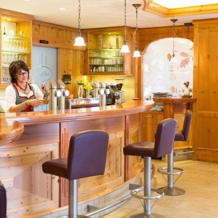 Eine Frau steht hinter einer Bartheke in einem rustikalen Raum mit Holzwänden, Regalen und Barhockern.