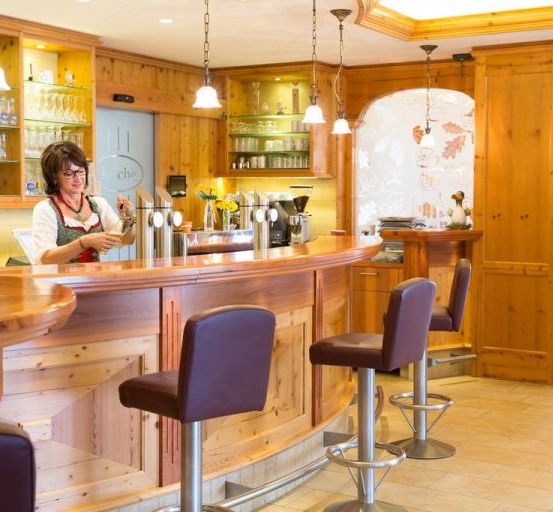 Eine Frau steht hinter einer Bartheke in einem rustikalen Raum mit Holzwänden, Regalen und Barhockern.