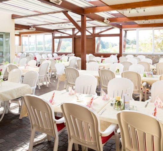 Ein Außenbereich zum Essen mit Tischen und Stühlen, geschmückt mit Tischdecken und Blumen. Die Decke besteht aus Metallplatten und die Wände aus Glas.