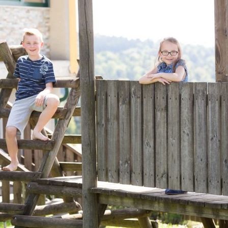 Zwei Kinder sind auf einem Holzspielhaus mit Blick auf einen Hügel. Der Junge ist barfuß auf der Leiter und das Mädchen lehnt sich an das Geländer.