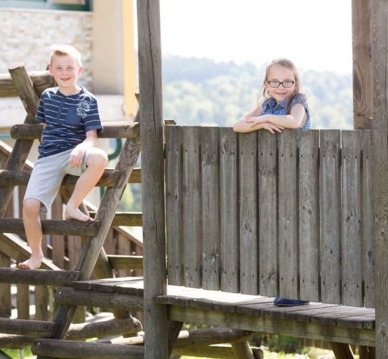 Zwei Kinder sind auf einem Holzspielhaus mit Blick auf einen Hügel. Der Junge ist barfuß auf der Leiter und das Mädchen lehnt sich an das Geländer.