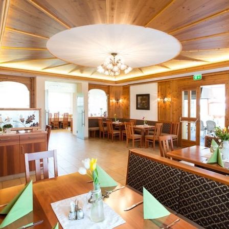 Ein Restaurantinnenraum mit Holzwänden und -decke. Eine Kronleuchter hängt von der Decke. Tische und Stühle sind aufgestellt, und es gibt Blumen und Servietten auf den Tischen.