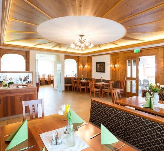 Ein Restaurantinnenraum mit Holzwänden und -decke. Eine Kronleuchter hängt von der Decke. Tische und Stühle sind aufgestellt, und es gibt Blumen und Servietten auf den Tischen.