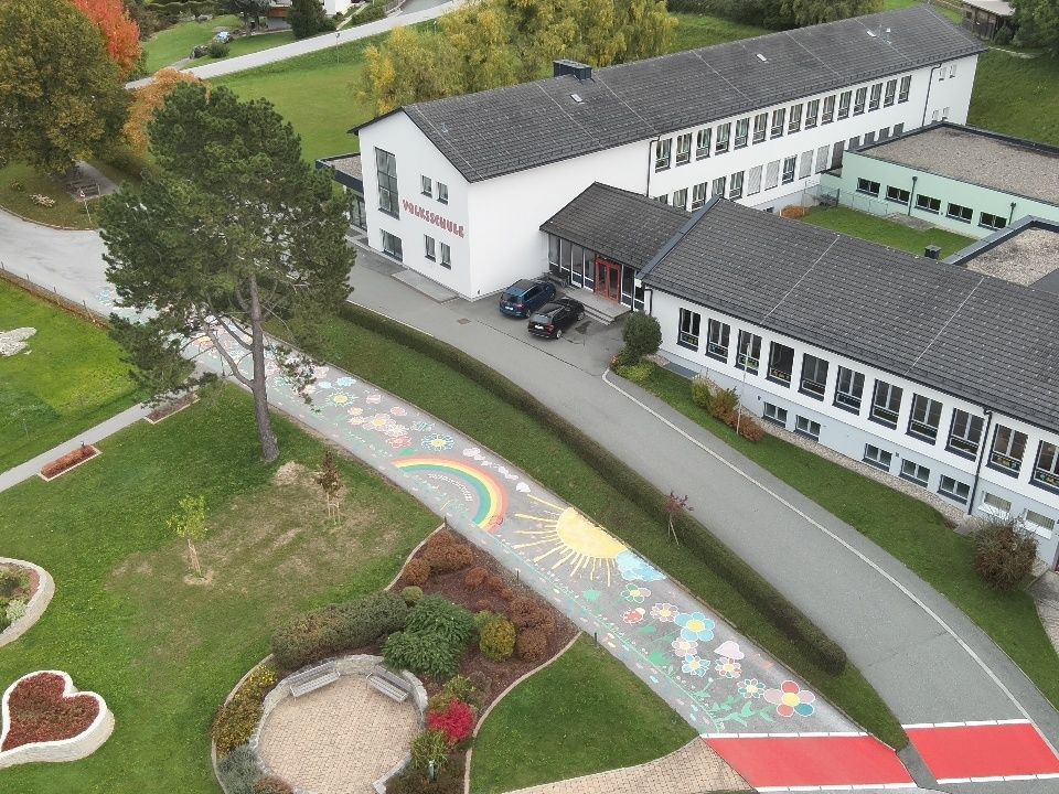 Eine Draufsicht auf eine Schule mit einem Regenbogen und einer Sonne auf dem Weg. Zwei Autos sind vor dem Gebäude geparkt.
