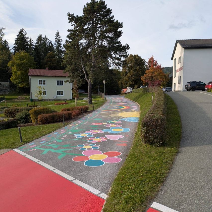 Ein bunter Fußweg mit gemalten Blumen und Herzen führt zu einem weißen Haus mit rotem Dach und einem großen Baum davor.