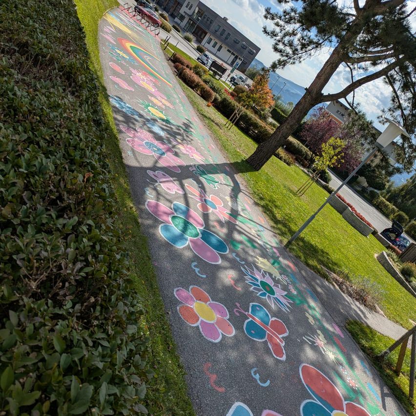 Ein buntes Kreide-Bild mit Blumen und Schmetterlingen ziert einen Weg im Park, mit einem Baum und Büschen an der Seite. Gebäude und geparkte Autos sind in der Ferne zu sehen.