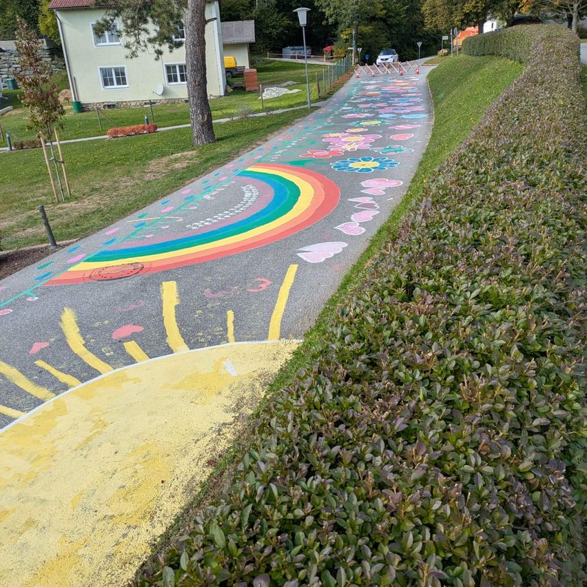 Ein bemalter Weg zeigt einen großen Regenbogen, Blumen und eine Sonne, mit einem grünen Haus und Autos im Hintergrund.