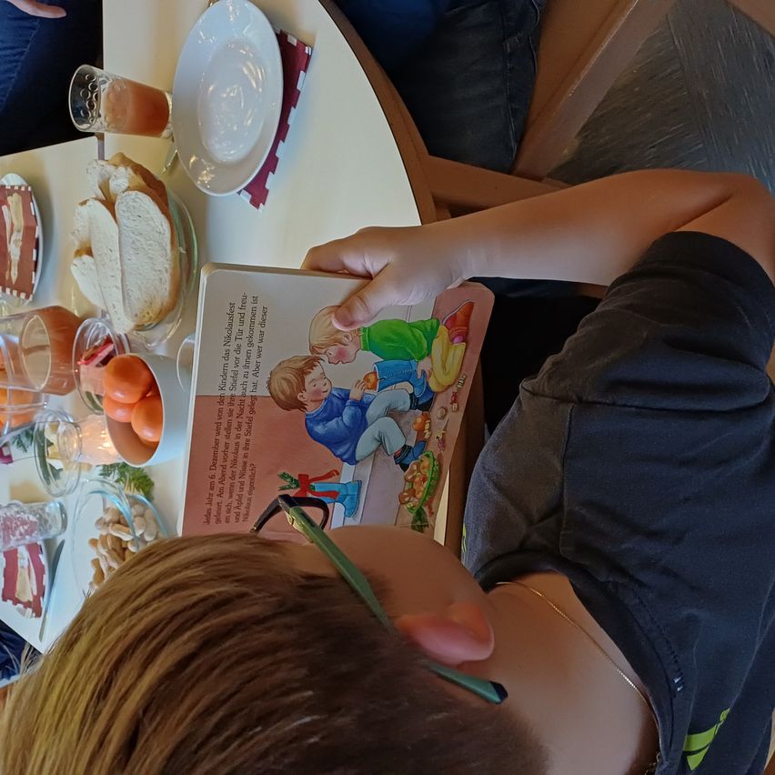 Ein Kind sitzt am Tisch und liest ein Buch. Der Tisch ist mit Essen gedeckt, darunter Brot, Eier und Saft. Eine andere Person sitzt in der Nähe.