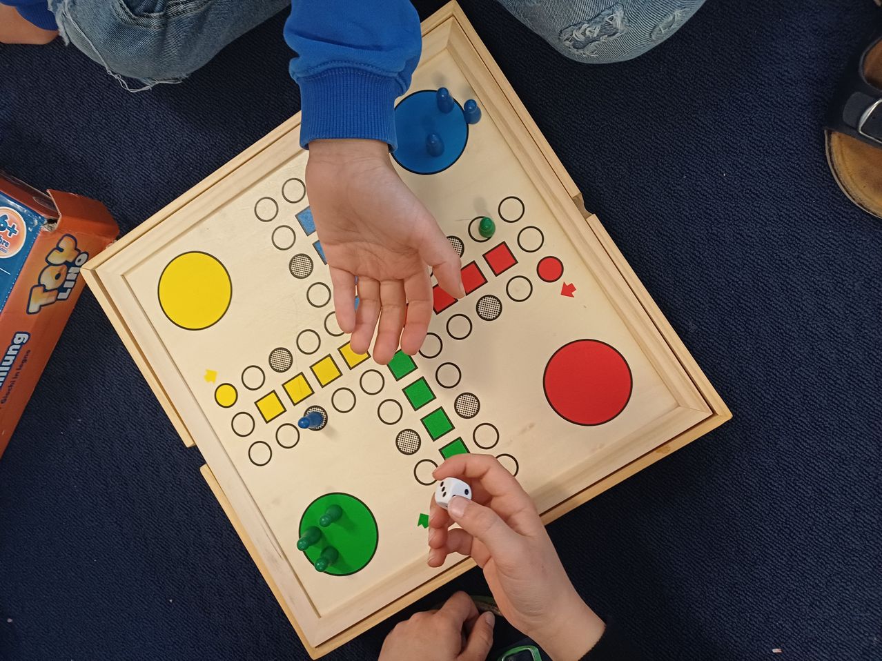 Zwei Personen spielen ein Brettspiel mit einem Würfel. Das Brett ist hölzern mit verschiedenen farbigen Teilen und einem Gitter. Ein Spieler würfelt.